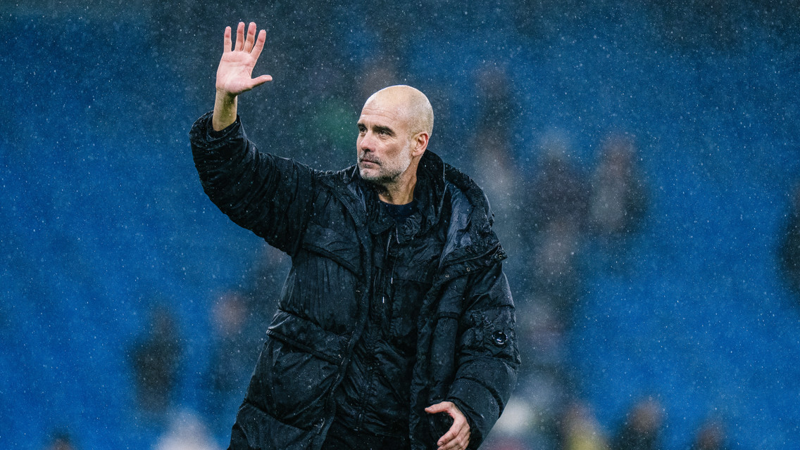 A person wearing a black padded jacket waves on a rain-soaked football field with a blurred face.