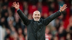 A man with a bald head and beard is wearing a black jacket. He is raising his hands and smiling in a celebratory manner against a blurred background of a crowd.