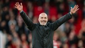 A man with a bald head and beard is wearing a black jacket. He is raising his hands and smiling in a celebratory manner against a blurred background of a crowd.