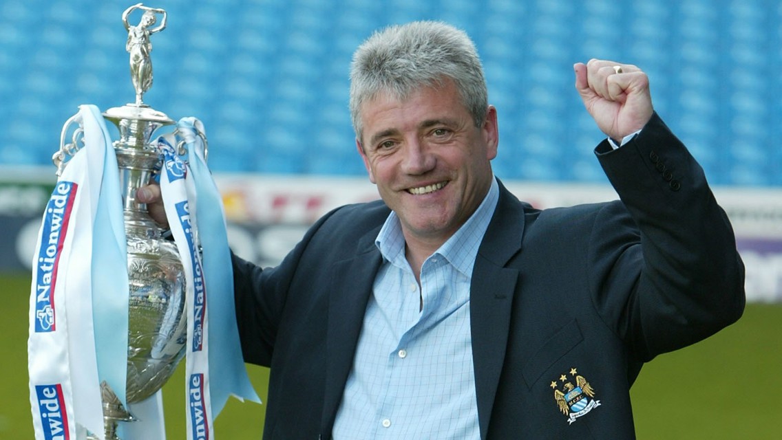 A person holding a trophy with Manchester City logo on their blazer, celebrating with a raised fist.