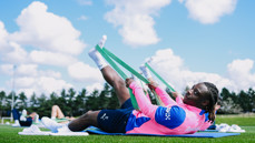 Athletes in pink jerseys use resistance bands for stretching exercises on a grassy field.