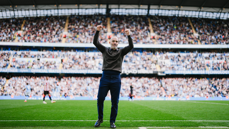 A man in a stadium stands on the football pitch with raised fists, celebrating. The stands are filled with spectators.
