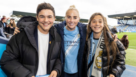 A Manchester City player in a home jersey poses with two smiling fans at a football stadium. The player is in the center, while the fans hold autographs and wear team merchandise.