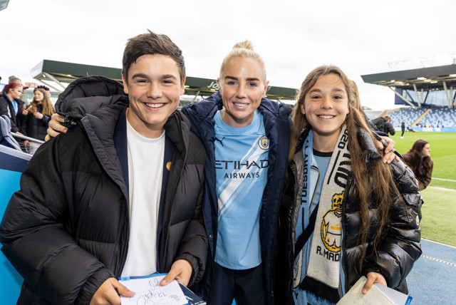A Manchester City player in a home jersey poses with two smiling fans at a football stadium. The player is in the center, while the fans hold autographs and wear team merchandise.