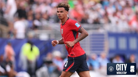A football player wearing a red and navy jersey with number 3 is running on the field. The background shows a blurred crowd, and there's a CITY AT THE WORLD CUP logo.