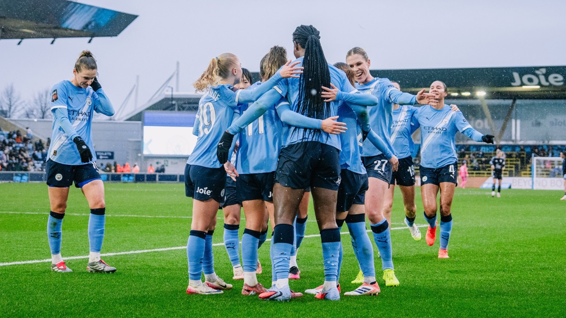 Manchester City women's team players celebrating on the pitch. They are wearing blue jerseys with 'Etihad Airways' and 'Joie' logos, in a group hug after a successful play.