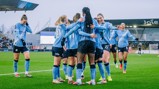 Manchester City women's team players celebrating on the pitch. They are wearing blue jerseys with 'Etihad Airways' and 'Joie' logos, in a group hug after a successful play.