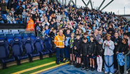 Crowd of fans at a stadium with empty seats visible, many people are standing behind barriers, an organizer in an orange jacket is present.