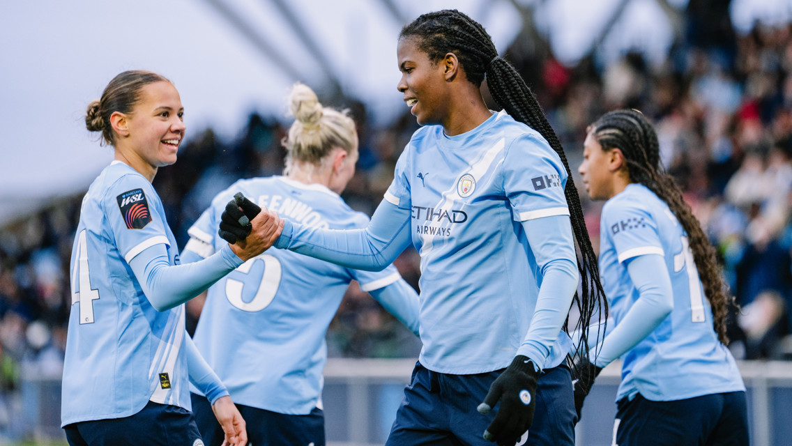 Four Manchester City players in light blue uniforms are celebrating on a soccer field. Two players are shaking hands.