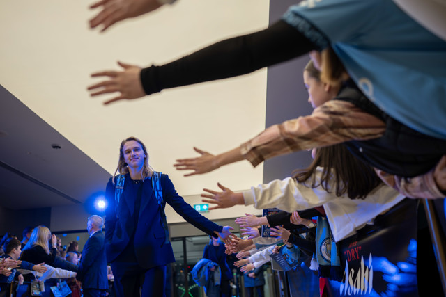 A person with a blurred face walks through a corridor lined with fans in a stadium or event setting, reaching out for high-fives or handshakes.