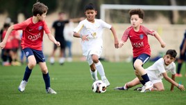 Children playing soccer on a green field. The players are wearing jerseys, some red with a logo and others white. A child in a white jersey is dribbling the ball.