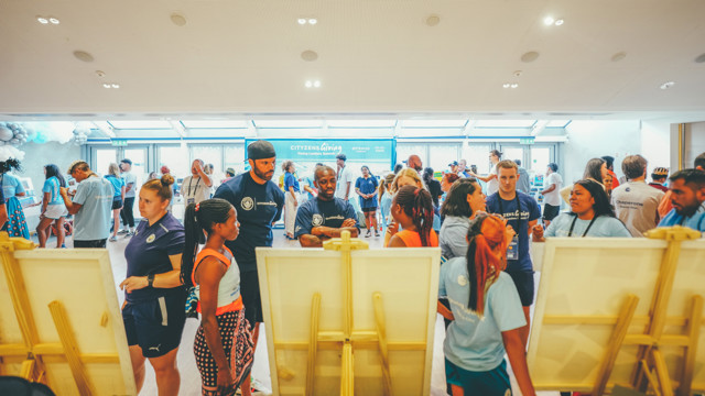 People of various ages are gathered at an indoor event. Multiple individuals wear 'Cityzens Giving' shirts. The room is well-lit with a banner in the background.