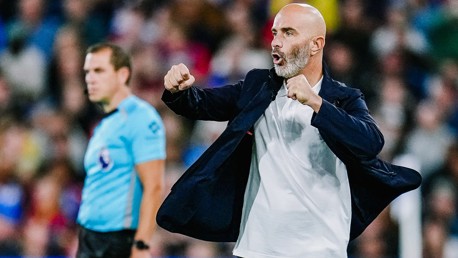 A soccer coach wearing a dark blazer and white shirt, making a dynamic gesture on the sideline during a match. An official in blue is visible in the background.