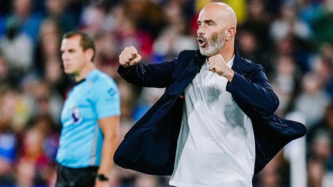 A soccer coach wearing a dark blazer and white shirt, making a dynamic gesture on the sideline during a match. An official in blue is visible in the background.