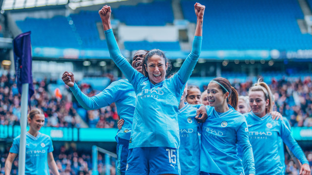 Manchester City Women's team players celebrating on the field in blue jerseys after a goal.