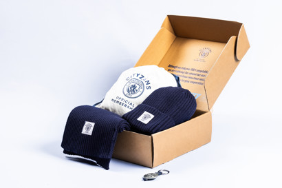 A membership pack with Manchester City branded items including a white drawstring bag, navy blue scarf and beanie with 'Cityzens Official Membership' logo in an open cardboard box.