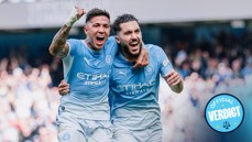 Two football players in Manchester City jerseys celebrate a goal on the field. The emblem is visible on their shirts.