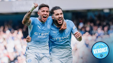 Two football players in Manchester City jerseys celebrate a goal on the field. The emblem is visible on their shirts.