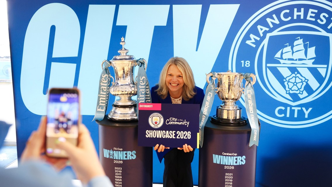 A person holding a 'Showcase 2026' sign stands between the Emirates FA Cup and Carabao Cup trophies at a Manchester City event.