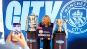 A person holding a 'Showcase 2026' sign stands between the Emirates FA Cup and Carabao Cup trophies at a Manchester City event.
