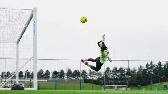 A goalkeeper in mid-air attempting to save a ball during soccer practice at a sports field.