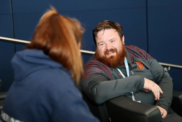 A man and woman are engaged in a conversation. The face of the man is blurred. The background has a dark blue wall.