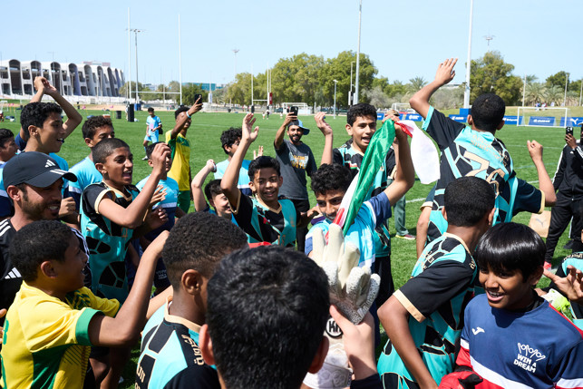 A group of young footballers celebrating on a football field, with arms raised and phones capturing the moment.