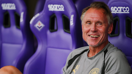 A man wearing a Manchester City shirt is smiling while sitting on a bench with purple Sparco seats.
