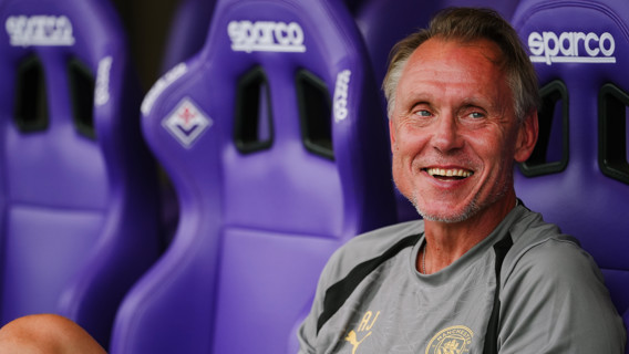 A man wearing a Manchester City shirt is smiling while sitting on a bench with purple Sparco seats.