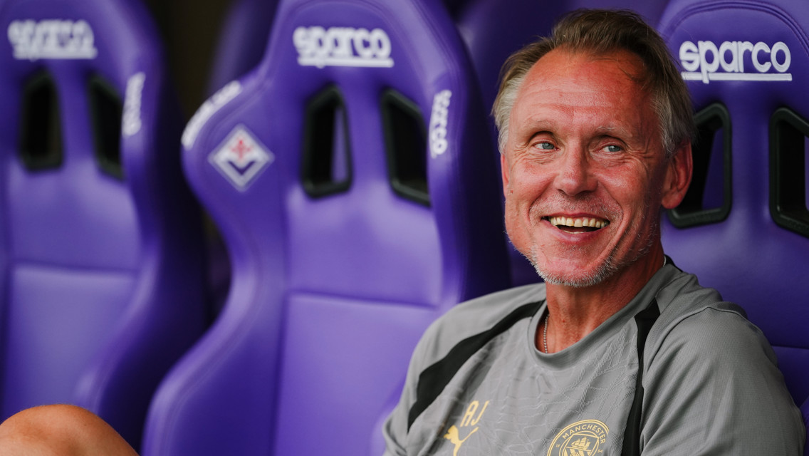 A man wearing a Manchester City shirt is smiling while sitting on a bench with purple Sparco seats.