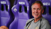 A man wearing a Manchester City shirt is smiling while sitting on a bench with purple Sparco seats.