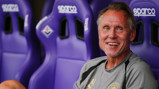 A man wearing a Manchester City shirt is smiling while sitting on a bench with purple Sparco seats.