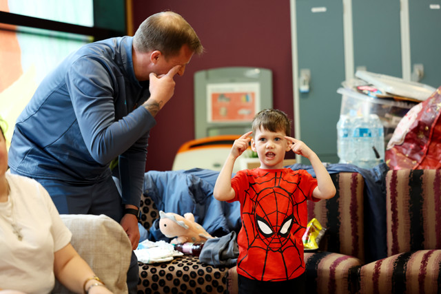 An adult in a blue tracksuit interacts with a child wearing a red Spiderman T-shirt, posing and pointing to their heads in a playful setting with toys and water bottles in the background.