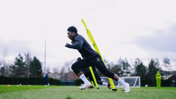 An athlete is sprinting on a football training field, near agility poles, with other equipment visible in the background.