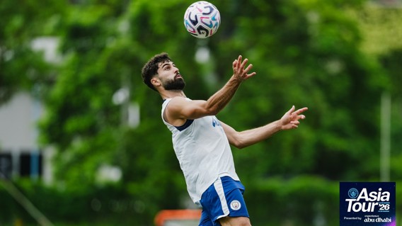 A soccer player is training outdoors, wearing a sleeveless shirt and shorts. The background is green with trees. The text 'Asia Tour '26 presented by Abu Dhabi' is displayed in the corner.