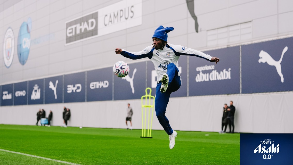 A soccer player in a Manchester City training session kicks a ball mid-air on a grassy pitch, wearing a blue hat and team tracksuit. Advertisements for Etihad and Puma visible in the background.