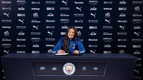 A person in a Manchester City jacket is signing a contract at a table with the club's logo. The background displays multiple sponsor logos.