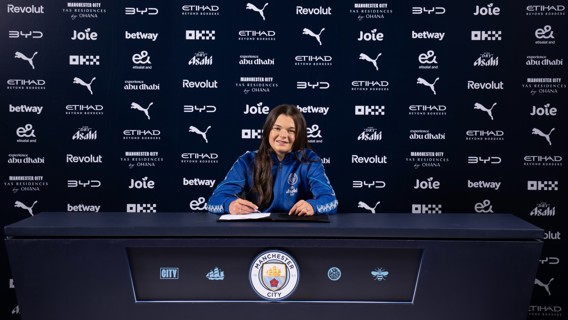 A person in a Manchester City jacket is signing a contract at a table with the club's logo. The background displays multiple sponsor logos.