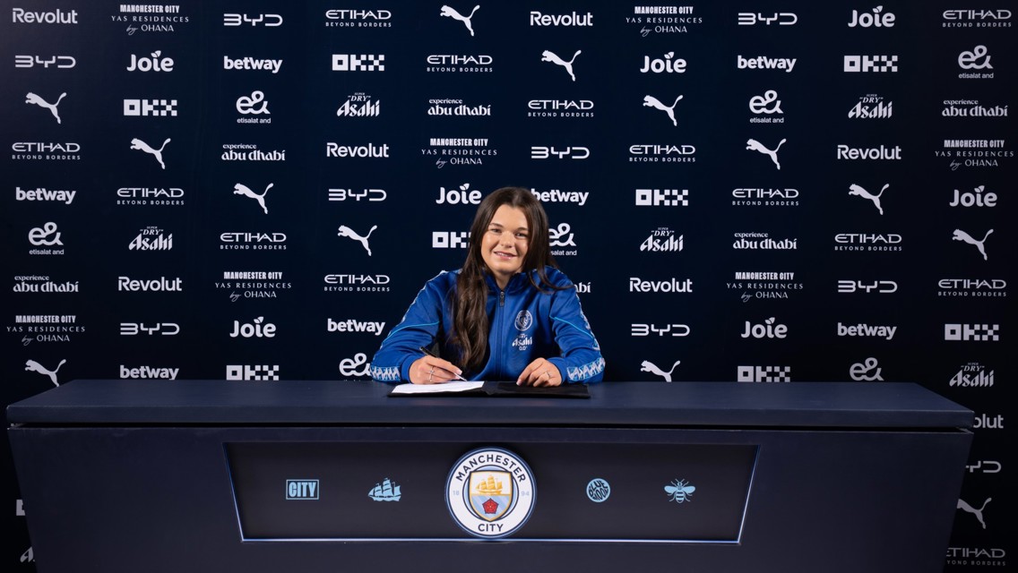 A person in a Manchester City jacket is signing a contract at a table with the club's logo. The background displays multiple sponsor logos.