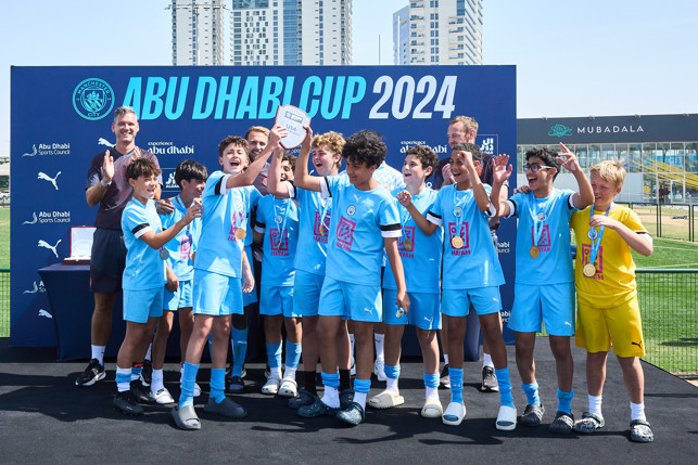 Group of young boys in light blue football kit celebrating their victory at Abu Dhabi Cup 2024 with medals and trophy. They stand in front of a banner with the event details.