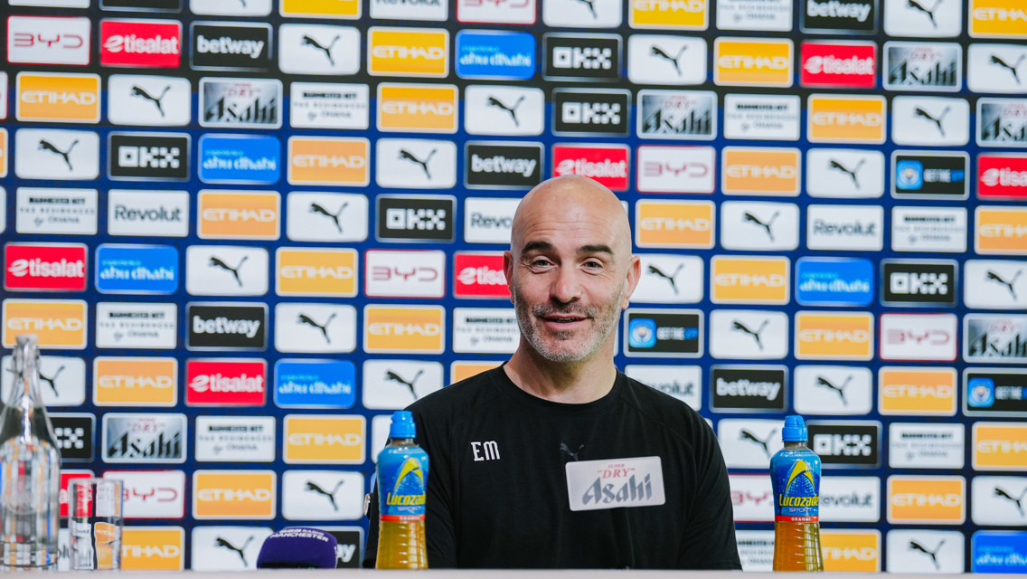 A man at a press conference table with branded sponsor backdrop. Items like water bottles and microphones are visible on the table.