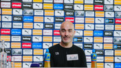 A man at a press conference table with branded sponsor backdrop. Items like water bottles and microphones are visible on the table.