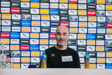 A man at a press conference table with branded sponsor backdrop. Items like water bottles and microphones are visible on the table.