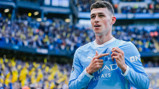 A soccer player in a Manchester City uniform points to the team crest on his jersey amid a stadium crowd.
