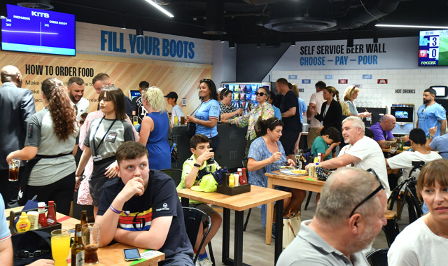 A crowded restaurant with people dining and ordering food, screens displaying order status, and a self-service beer wall.
