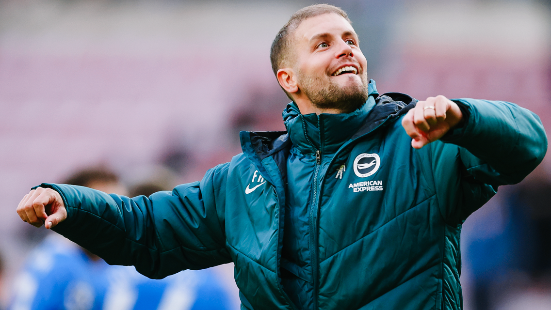 A person wearing a Brighton & Hove Albion jacket is celebrating, with arms raised and a joyful expression.