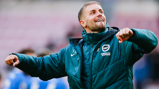 A person wearing a Brighton & Hove Albion jacket is celebrating, with arms raised and a joyful expression.