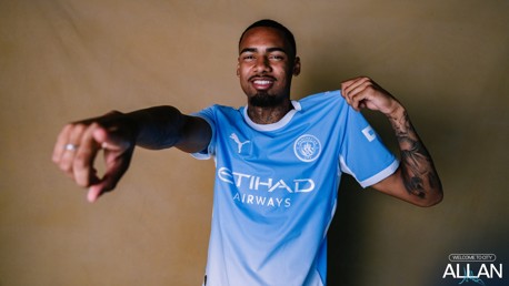 A person wearing a Manchester City jersey, pointing forward and holding the shirt collar with a smile.