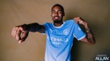 A person wearing a Manchester City jersey, pointing forward and holding the shirt collar with a smile.