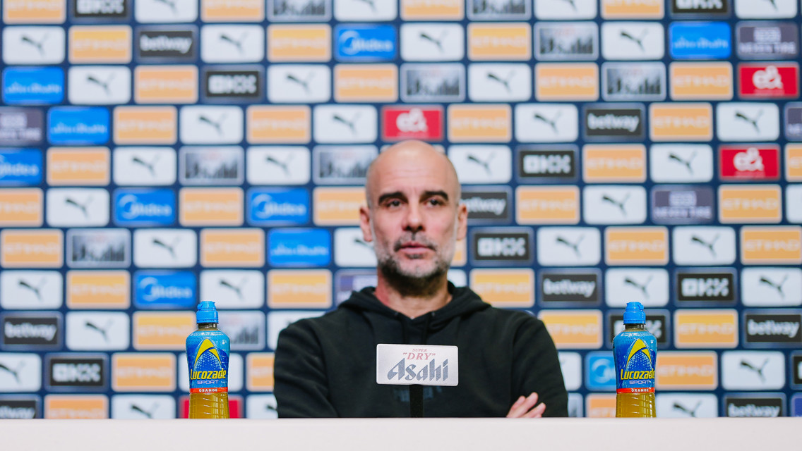 A press conference setting with a blurred individual, featuring a sponsor background wall with Puma and Lucozade branding.